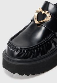Juodai lakoti loaferiai su banguotu kraštu, su širdelės formos auksiniu akcentu ant dirželio ir storu, tekstūruotu juodu padu.