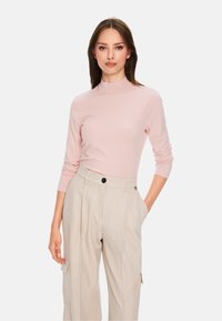 Hellrosa, figurbetontes Langarmoberteil mit hohem Kragen, kombiniert mit beigefarbenen, weit geschnittenen Hosen, die über einen schwarzen Knopf und seitliche Taschen verfügen.