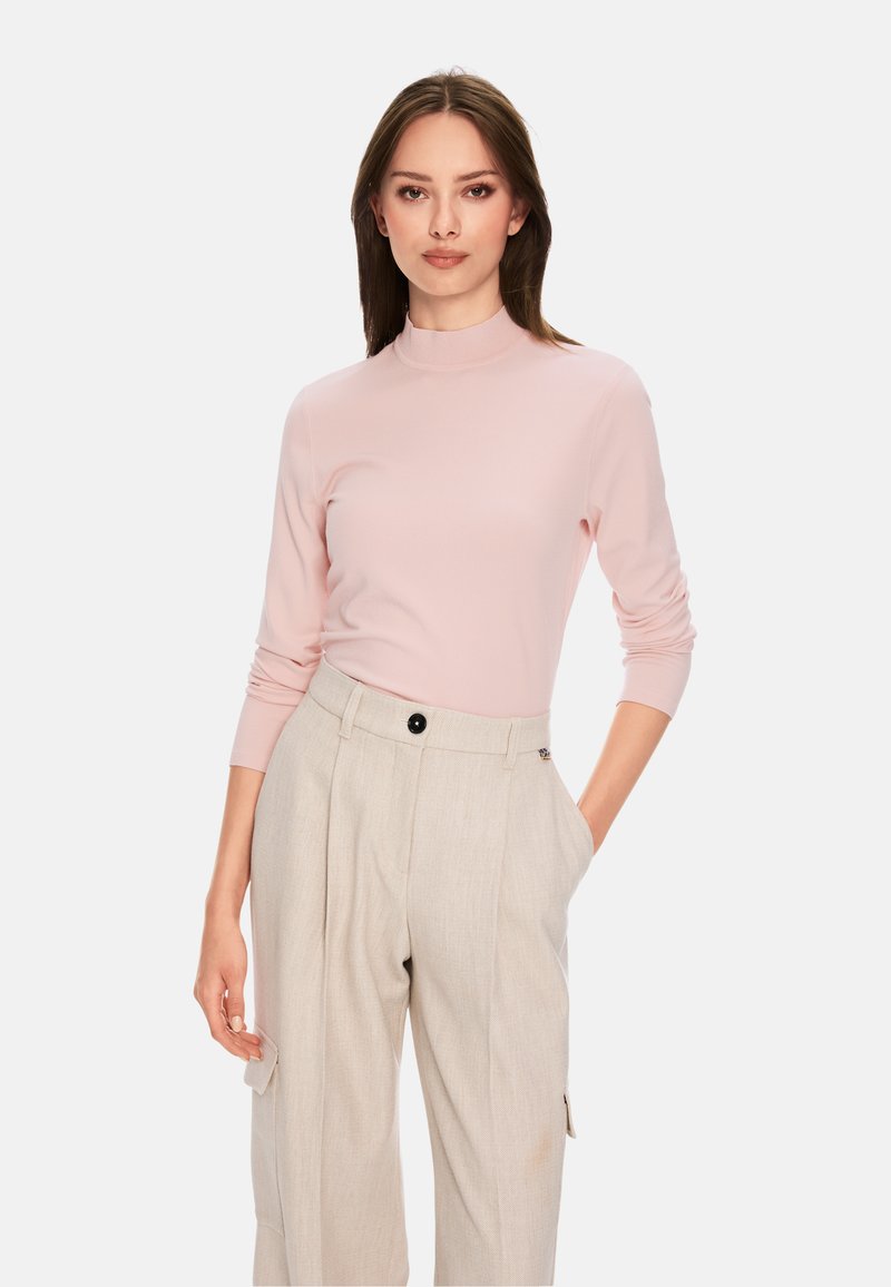 Hellrosa, figurbetontes Langarmoberteil mit hohem Kragen, kombiniert mit beigefarbenen, weit geschnittenen Hosen, die über einen schwarzen Knopf und seitliche Taschen verfügen.