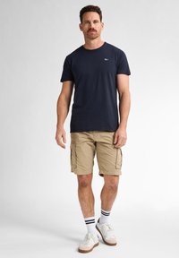 Marineblauw korte mouwen t-shirt, kaki cargoshorts met zijzakken, witte sneakers en gestreepte crew-sokken, staande tegen een effen achtergrond.