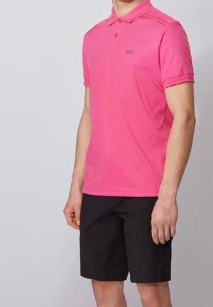 Mann trägt ein leuchtend pinkes kurzärmeliges Polohemd und schwarze knielange Shorts, steht vor einem schlichten grauen Hintergrund.