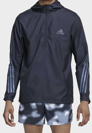 Mężczyzna ubrany w granatową bluzę z kapturem Adidas z trzema paskami na rękawach oraz szare i czarne krótkie spodenki w kamuflażu Adidas.