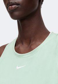 Κοντινό πλάνο ενός ατόμου που φοράει αμάνικο τοπ σε μέντα με λευκό λογότυπο Nike στο στήθος, σε απλό ανοιχτό φόντο.