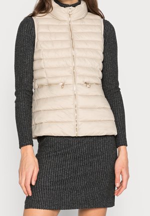 Vrouw draagt een beige doorgestikte mouwloze vest met ritssluitingen over een donkergrijze geribbelde jurk met lange mouwen.