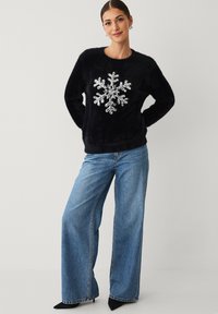 Zwarte, pluizige trui met een zilveren sneeuwvlokontwerp in het midden, gecombineerd met wijde blauwe spijkerbroek en zwarte puntige schoenen.