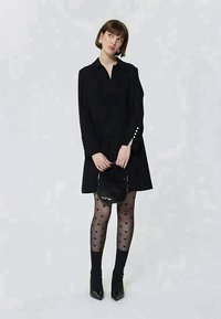 Femme dans une robe noire à manches longues, collants transparents à motif cœur, escarpins noirs à bout pointu, tenant un petit sac à main noir, se tenant devant un décor uni.