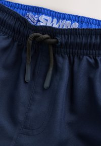 Tmavomodré plavkové šortky s modrým elastickým pásom, vybavené sťahovacou šnúrkou a hladkou textúrou látky. Malé logo v hornej časti.