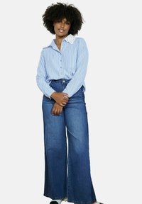 Zila un balta svītraina pogājama krekla ar mežģīņu apkakli kombinācija ar plašiem tumša džinsa biksēm. Modelis stāv ar rokām krustām.