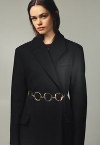 Ung kvinde med blå øjne iført en oversize sort blazer, der er spændt ind med et gyldent kæde bælte mod en ensartet grå baggrund.