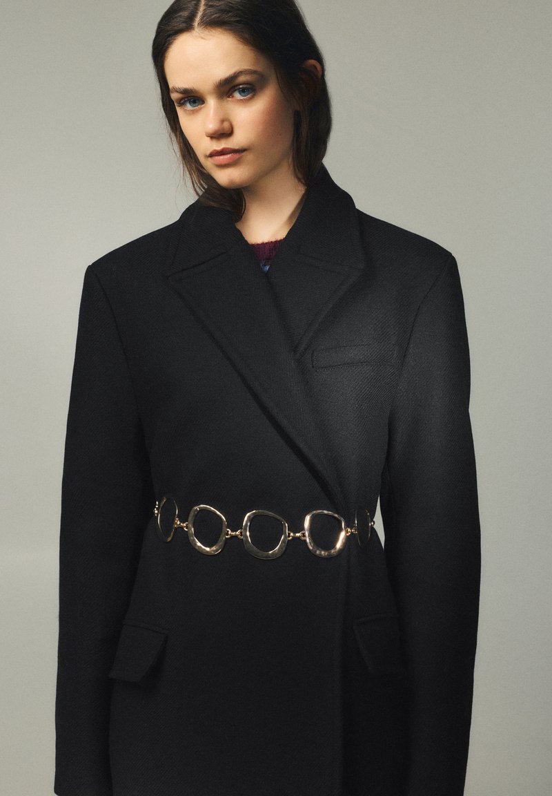 Ung kvinde med blå øjne iført en oversize sort blazer, der er spændt ind med et gyldent kæde bælte mod en ensartet grå baggrund.