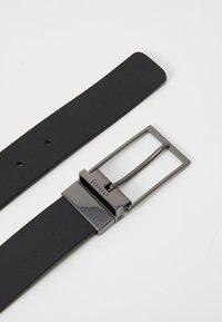 Ceinture en cuir noir avec une boucle rectangulaire en métal, affichant une finition polie et un logo de marque gravé sur la boucle.