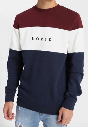 Henkilö pukeutuneena värilohkoneuleeseen, jossa on viininpunaisia, valkoisia ja laivastonsinisiä raitoja ja rinnan kohdalla kirjailtu sana "BORED".