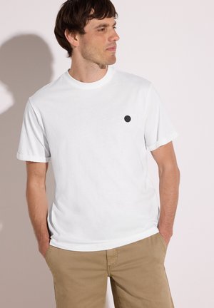 Mand står med hænderne i lommerne iført en ensfarvet hvid kortærmet t-shirt med en lille sort cirkel på brystet og khaki bukser.