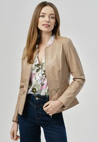 Skórzana kurtka w kolorze brązowym o dopasowanym kroju, z zamykanymi kieszeniami i stojącym kołnierzem, noszona na bluzce w kwiatowy wzór i ciemnych dżinsach.