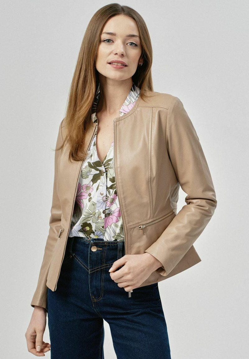 Skórzana kurtka w kolorze brązowym o dopasowanym kroju, z zamykanymi kieszeniami i stojącym kołnierzem, noszona na bluzce w kwiatowy wzór i ciemnych dżinsach.