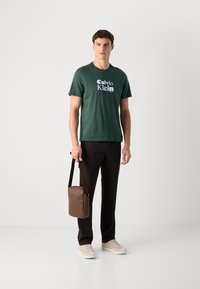 Man standing wearing a green Calvin Klein t-shirt, black pants, beige sneakers, and holding a brown leather shoulder bag against a white background.