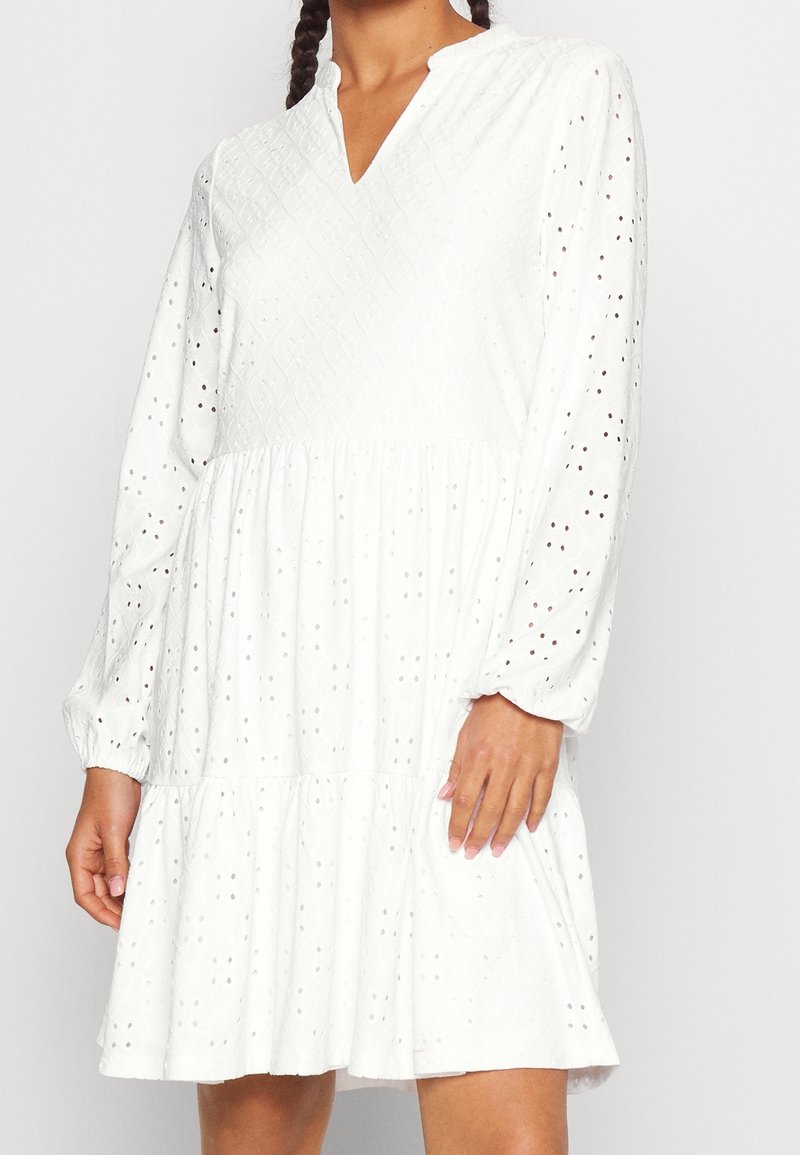 Femme portant une robe blanche à manches longues en tissu ajouré avec un col en V, debout devant un fond clair uni.