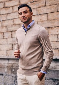 Beige ritsneck sweater met een zachte textuur, gedragen over een lichtblauw overhemd met kraag. Model staat tegen een bakstenen muur met de handen in de zakken.