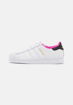 Biały sneaker Adidas Superstar z różową podszewką, czarną wstawką z tyłu, złotym logo i trzema białymi paskami po boku.