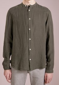 Olivově zelená lněná košile s dlouhými rukávy, vysokým límcem a bílými knoflíky, se uvolněným střihem a zaobleným spodním lemem.