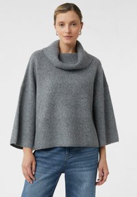 Femme portant un pull gris loose à côtes avec un large col châle et des manches mi-longues, associé à un jean bleu, se tenant contre un fond uni.