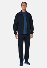 Giacca con zip blu navy con bordi bianchi, abbinata a una t-shirt blu e pantaloni blu navy. L'outfit presenta un design sportivo e una vestibilità comoda.