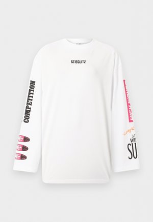 Vrouw draagt een wit shirt met lange mouwen met zwarte en rode tekst, grijze wijde broek, zwarte schoenen en een zwarte pet, staand met handen in de zakken.