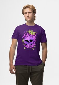 Fialové bavlněné tričko s grafickým designem lebky obklopené živými růžovými a zelenými graffiti prvky.