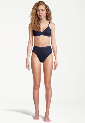 Giovane donna con capelli ricci, che indossa un reggiseno blu navy e mutande a vita alta, in piedi a piedi nudi davanti a uno sfondo bianco semplice.