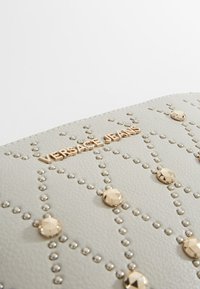 Sac à main en cuir gris clair texturé avec des clous dorés et le logo "VERSACE JEANS" embossé. Présente un motif géométrique avec des accents argentés.