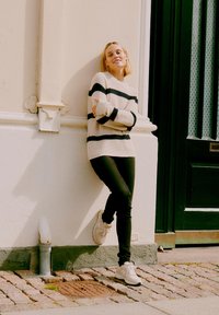 Strikket hvit genser med svarte striper, svarte leggings og hvite joggesko. Modellen lener seg mot en vegg med steinbasis og grønn dør.