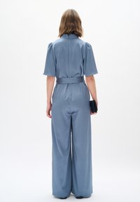 Blauer Jumpsuit mit tailliertem Bund, kurzen Puffärmeln und weitem Bein. Glatte, glänzende Textur mit einer einfarbigen Optik. Schwarze Clutch in der Hand.