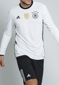 Vit långärmad sporttröja med svarta detaljer, prydd med en gyllene FIFA-emblem, fem stjärnor över logotypen och en tre-streksdesign på ärmarna.