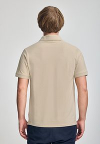Beżowa koszulka polo o gładkiej fakturze, z krótkimi rękawami i klasycznym kołnierzykiem, pokazana z tyłu. Prosty design, bez widocznych logo czy wzorów.