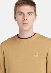 Lichtbruine gebreide trui met een geribbelde halslijn en een klein, vierkant Timberland-logo op de borst. Soepele textuur, lange mouwen.