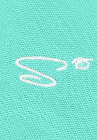 Türkiser Stoff mit strukturierter Webart, der ein auffälliges weißes gesticktes "S" und geschwungene Designelemente zeigt.