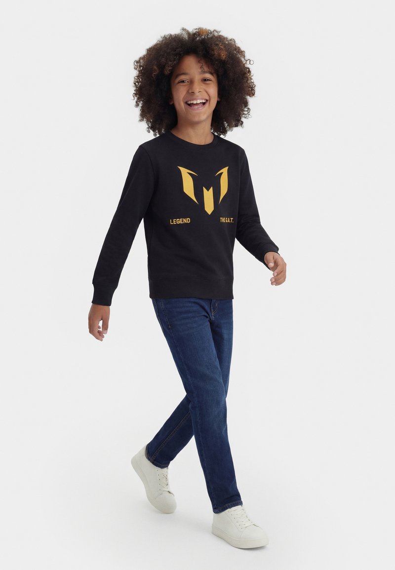 Enfant souriant aux cheveux bouclés marchant, portant un sweat-shirt noir avec un motif doré et du texte, un jean bleu et des baskets blanches sur un fond uni.