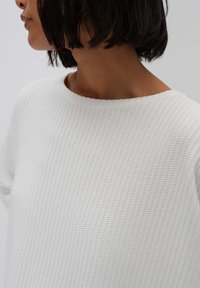 White textured sweatshirt with a round neckline, featuring a subtle ribbed pattern. The fabric appears soft and lightweight.