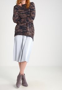 Osoba w brązowym i czarnym dzianinowym swetrze, białej plisowanej spódnicy midi i szarych zamszowych botkach na kostkę, stojąca na jednolitym tle.