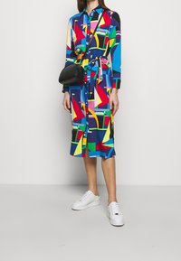 Färggjord klänning med en blandning av geometriska former i blått, rött, gult och grönt, med knyt i midjan och knappdesign. Bärs med vita sneakers.