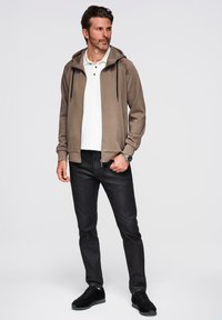 Sudadera cremoso con cremallera y capucha, cordones negros, llevada sobre una camiseta polo blanca. Jeans negros y zapatos negros completan el atuendo. Diseño simple.