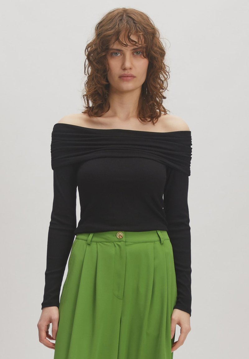 Zwarte off-shoulder top met een getextureerde, gedrapeerde halslijn; lange mouwen; gecombineerd met groen hoge taillebroeken met een knoop aan de voorkant.