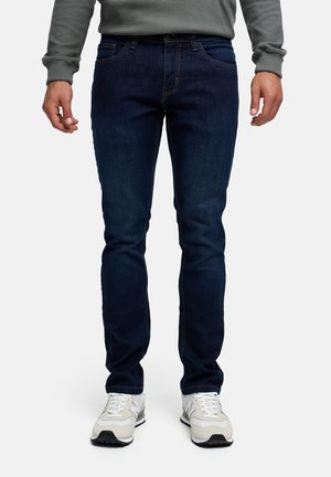 Mężczyzna ubrany w ciemnoniebieskie obcisłe dżinsy, szarą koszulę z długim rękawem i białe trampki, stojący prosto na jednolitym tle.