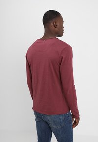 Langärmliges Shirt in Burgunderrot, aus strukturiertem Stoff, mit rundem Ausschnitt und abgerundetem Saum, sichtbare Nähte und ein Etikett.