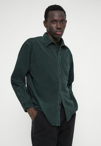 Camisa de côrdura verde escura com botões à frente, colarinho pontiagudo, bolso no peito e mangas longas, contra um fundo claro e liso.