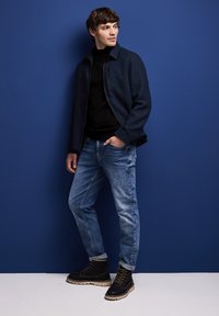 Marinblå jacka med hög krage, svart ribbad polotröja under, ljusblå jeans och svarta stövlar med gråa sulor, mot en blå bakgrund.