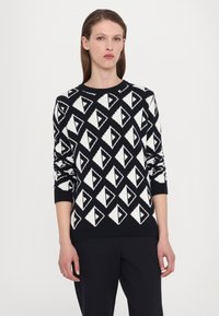 Svart og hvit genser med diamantmønster, strikket tekstur, rund hals, og ribbede mansjetter og kant. Designet er geometrisk.