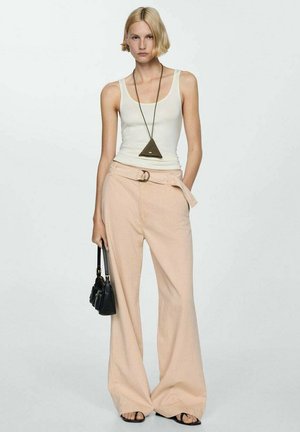 Pantaloni ampi color beige chiaro con cintura in vita, abbinati a un top bianco senza maniche e una collana con pendente triangolare; tiene in mano una piccola borsa nera.