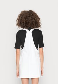 Vitt denimoverallklänning med korsade axelremmar, med en lös passform och en slät textur, bärs över en svart ribbad topp.