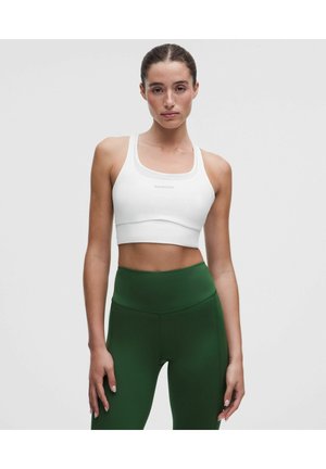 Vrouw met een witte sportbeha en donkergroene leggings met hoge taille, staand tegen een effen lichtgrijze achtergrond.